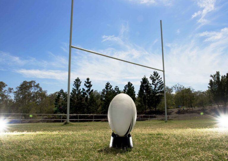 Rugby ball on a kicking tee in front of goalposts under a clear blue sky, symbolising teamwork and focus beyond the sidelines.
